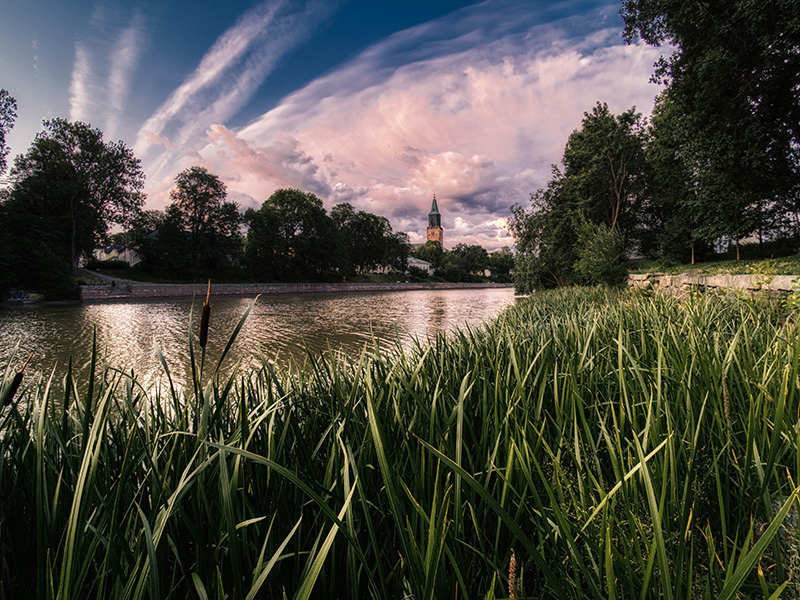 Turun Tuomiokirkko kohoaa horisontissa ja sen edessä virtaa Aurajoki. Taivaalla on upeat pilvimassat ja kuvan alareunassa vehreää ruohikkoa.