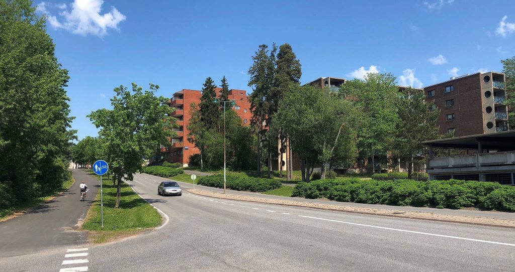 Turun Varissuon lähiön kerrostaloja ja rehevää puustoa. Etualalla tie, jossa kulkee auto ja tien reunassa pyöräilijä.