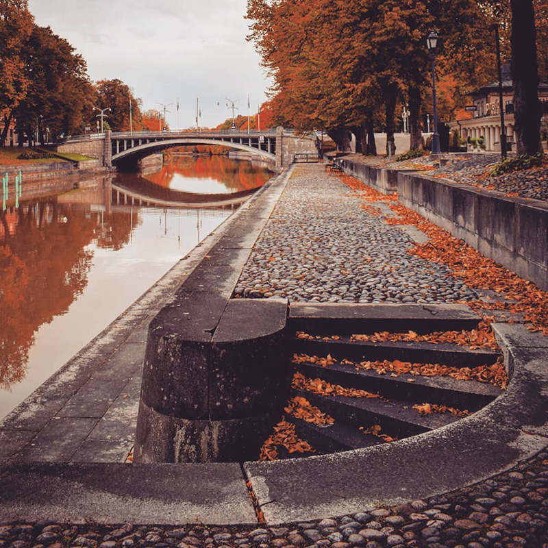 Turun keskustassa Aurajoen ranta värjäytyy oranssiksi syksyn lehdistä ja puiden latvoista. Aurajoen yli kulkee silta ja joen viertä tie mukulakivistä.
