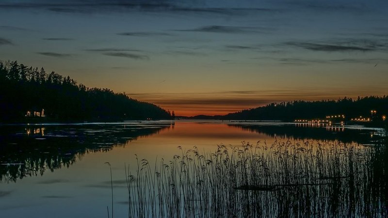 Auringonlasku Kuusistonsalmella häilyy oranssina hämärtyvän peilityynen merenpinnan yllä ja Kuusiston saarella ja Voivalan rannassa hohtaa talojen valoja.