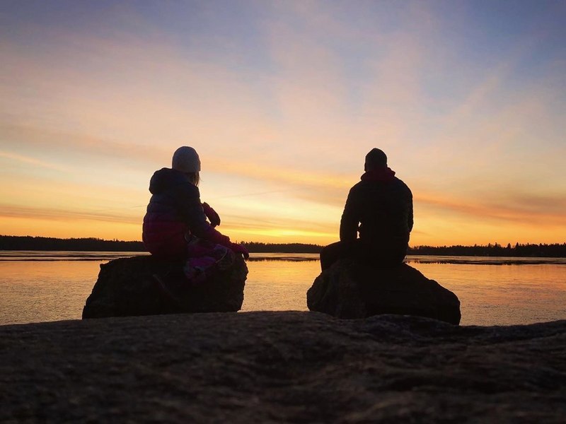 Kaksi tuulipukuista retkeilijää istuvat kallioisella rannalla kahden kiven päällä ihaillen auringonlaskun värjäämää oranssia taivasta.