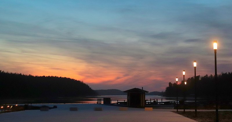Hovirinnan rannassa katuvalot valaisevat hämärtyvää iltaa, auringon viimeiset säteet värjäävät taivaanrannan oranssiksi ja taivaalla leijailee harsopilviä.