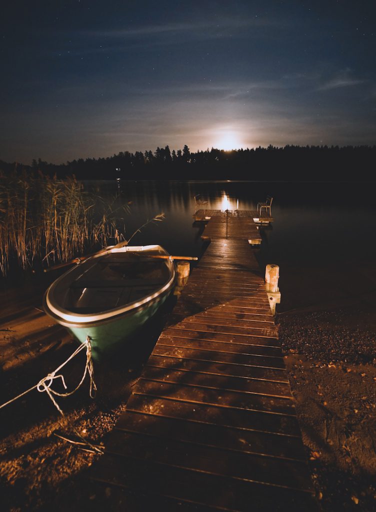 Soutuvene Kemiönsaaressa meren rannalla kiinnitettynä laituriin pimeän tullessa ja auringon viimesäteiden hohtaessa puiden siluetin takaa horisontissa.