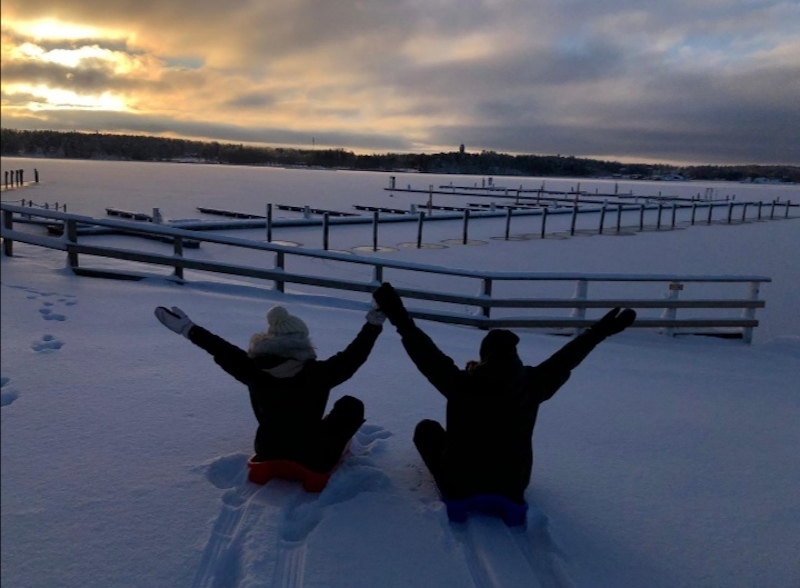 Kaksi pulkkailijaa istuvat pulkissa kädet ylhäällä ja katselevat jäätynyttä lumista merenrantaa.