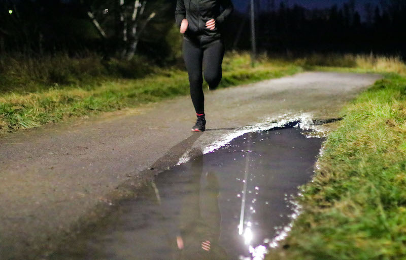 Lenkkeilijä juoksemassa katuvalojen loisteessa hiekkatiellä, jossa lätäköstä heijastuu katuvalo ja lenkkeilijän kuvajainen.