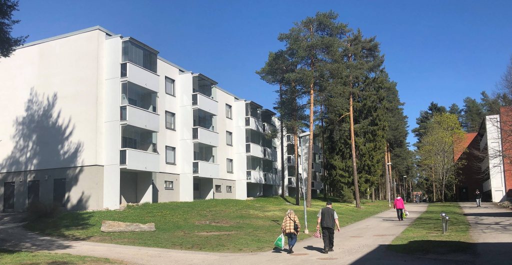 Jäkärlän ostoskeskuksen läheisyydessä oleva valkoinen kerrostalo ja muutamia ihmisiä kävelemässä kauppakassien kanssa sen edessä olevalla tiellä auringonpaisteessa.