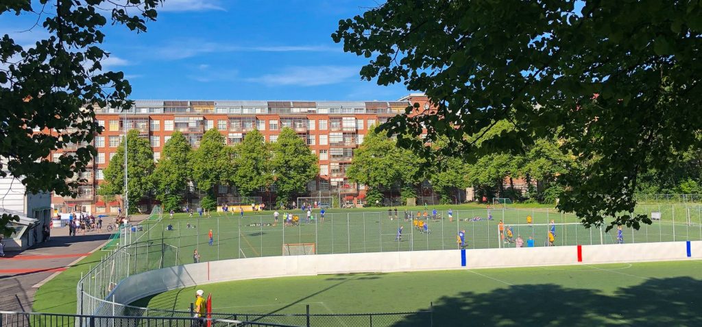 Turun Verkahovin punatiilinen kerrostalo taustalla ja Parkin jalkapallokenttä etualalla kesäisenä ja aurinkoisena päivänä. Kentällä useita pelaajia.