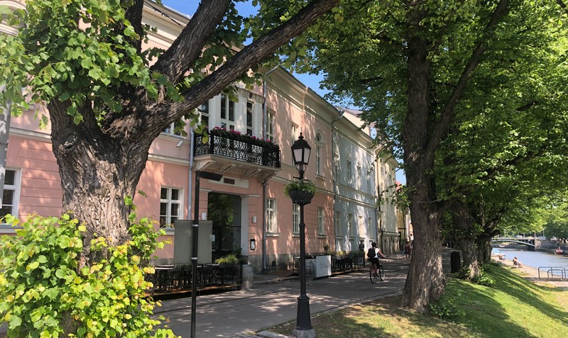 Aurajokirannassa kesällä vanha vaaleanpunainen rakennus, jossa parveke toisessa kerroksessa. Rannassa on myös lehmuksia, muutama ihminen ja pyöräilijä.