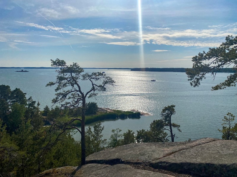 Maanpään kallioilta avautuva näkymä auringossa välkehtivälle merelle, jossa seilaa purjevene