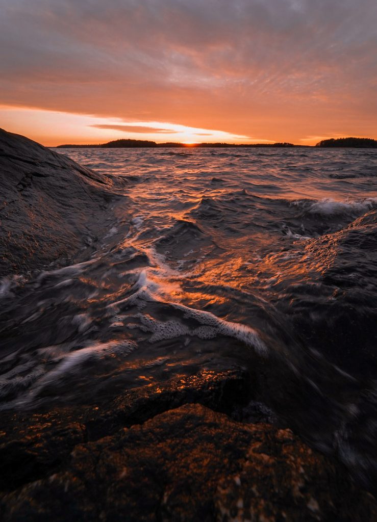 Kuohuavat meren rannan aallot ja niistä nousevat kalliot upean ilta-auringon värjääminä.