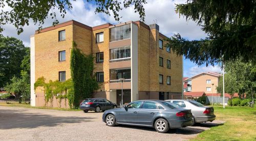 Vaalea kerrostalo Eurassa aurinkoisena päivänä, kolme pysäköityä autoa etualalla.