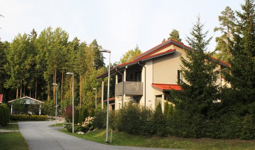 Vaalea omakotitalo kadun oikealla puolella. Puita ja pensaita etualalla. Metsää taustalla Tikanmaan asuinalueella Raisiossa.