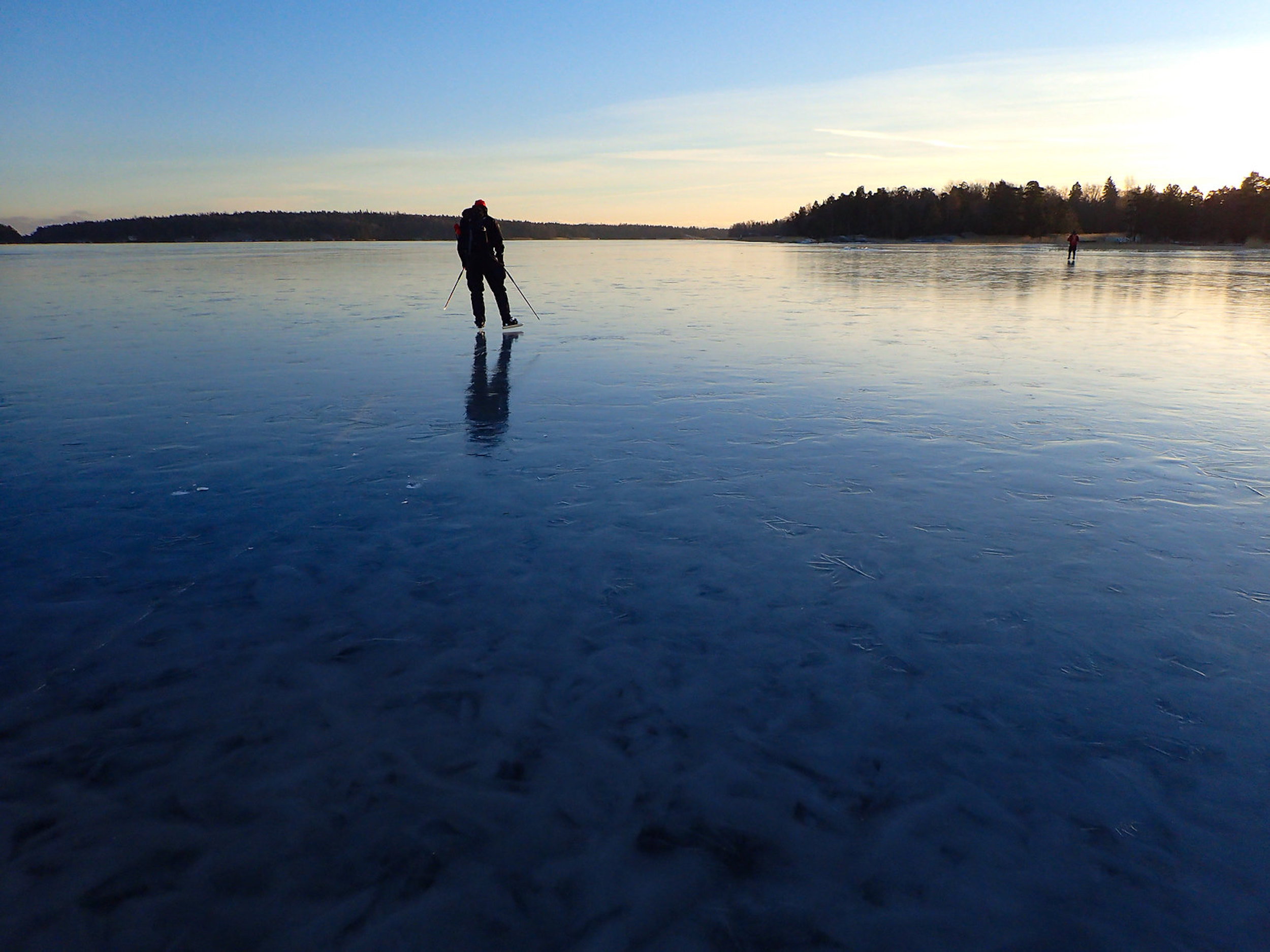 Retkiluistelija järven jäällä, taustalla laskevan auringon kajo.